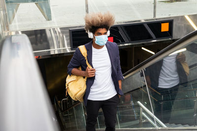 Young african man wearing a mask on an escalator with a bag hanging over his shoulder while listening to music with a wireless headset
