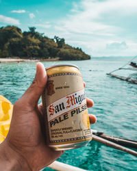 Close-up of hand holding drink at sea against sky