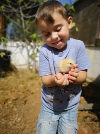 Cute smiling boy holding baby chicken on field during sunny day