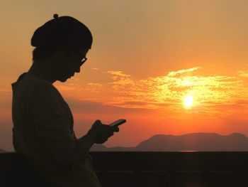 Side view of woman using phone against sky during sunset