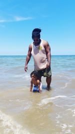 Father looking at son playing in water at beach against sky