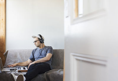 Man listening music while using laptop computer on sofa seen through doorway