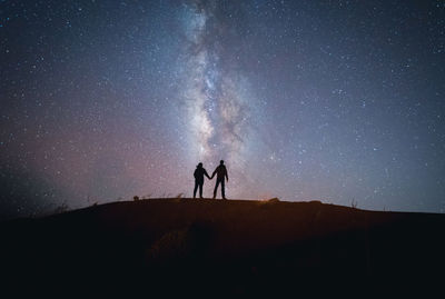 Back view of unrecognizable tender traveling couple standing on hill holding hands while admiring spectacular scenery of milky way over mountainous in starry night
