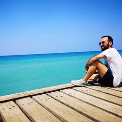 Man sitting by sea against clear blue sky