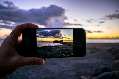 Cropped hand of person photographing sea on mobile phone against sky during sunset