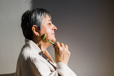 Side view of middle aged female in bathrobe massaging face with jade roller while standing with closed eyes on gray background during skincare procedure