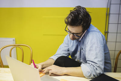 Young man using smart phone while writing on adhesive note at desk in board room