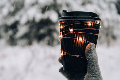 Female hand in winter warm gloves holding black eco paper cup with garland shiny light. creative 