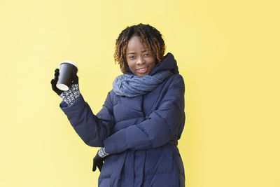Portrait of smiling young woman holding coffee cup against yellow background