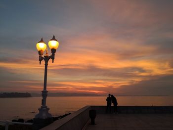 Man and illuminated street light by sea against sky at sunset