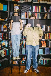 Male and female friends covering face with books while standing near shelf in bookstore