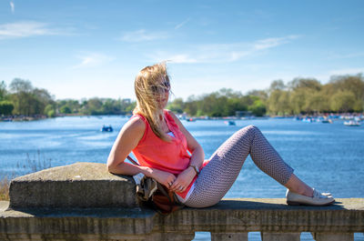 Woman with blond hair sitting on retaining wall against lake