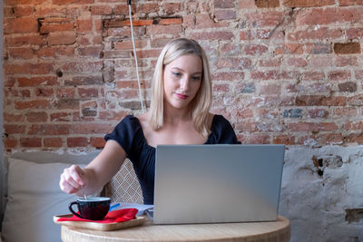 Young woman using laptop while standing against brick wall
