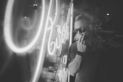 Young man wearing sunglasses and smoking while standing by illuminated lights at night