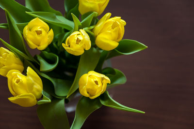 A bouquet of yellow tulips with green leaves on a brown board background, top view