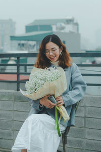 Young woman holding bouquet while sitting against wall in city