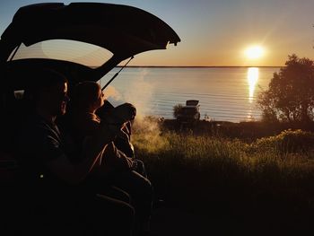 People sitting on land by lake against sky during sunset