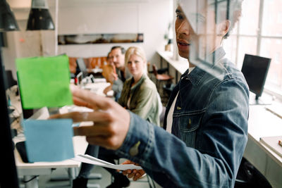 Businessman showing adhesive note to creative team seen from glass at workplace