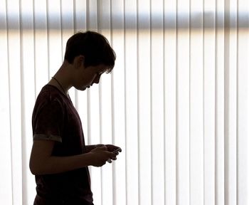 Side view of teenage girl texting by window blinds