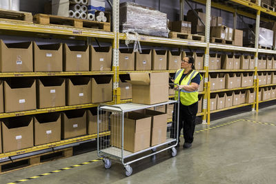 Full length of female worker pushing cart with cardboard boxes by rack at warehouse