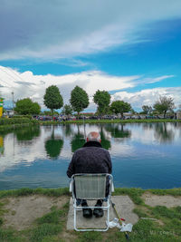 Rear view of man sitting by lake against sky