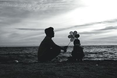 Father and son with pinwheel crouching on sea shore against sky