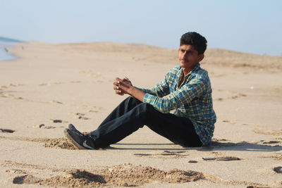 Young man sitting on sand at beach against clear sky