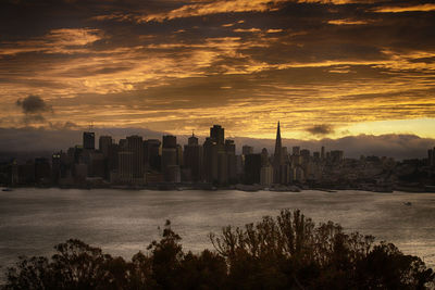 Urban skyline by sea against cloudy sky during sunset