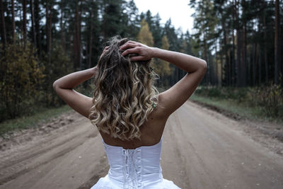 Rear view of young woman with hand in hair standing on road at forest