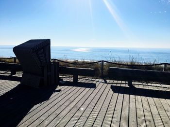 Wooden bench by sea against sky on sunny day