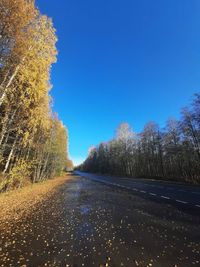 Empty road amidst trees against clear blue sky