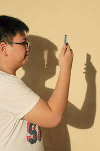 Side view of teenage boy using smart phone against beige colored wall