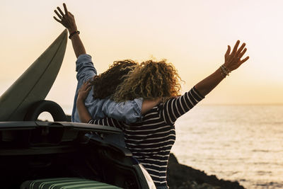 Rear view of friends with arms raised standing by car at beach against sky during sunset