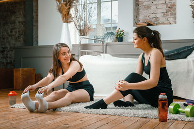 Two athletes arestretch in the bedroom, in the background there is a bed, a vase, a carpet.