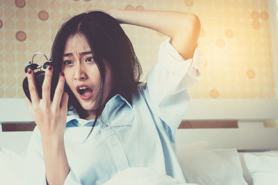 Shocked young woman holding alarm clock on bed at home