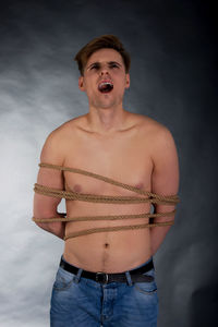 Shirtless young man tied with rope shouting against gray background