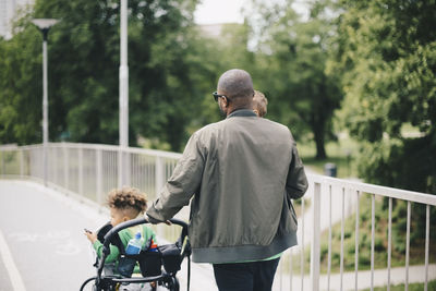 Rear view of father pushing baby stroller while walking with sons on bridge