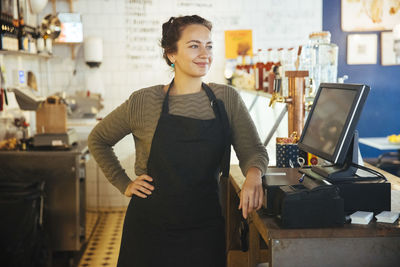 Confident young female owner standing with hand on hip by cash register at checkout in cafe