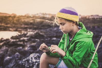 Boy sitting by fishing rod on rock against sky during sunset