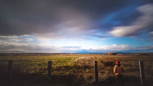 Rear view of man standing by fence on grassy field against cloudy sky