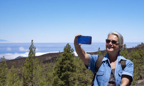 Portrait of smiling woman photographing through smart phone against blue sky