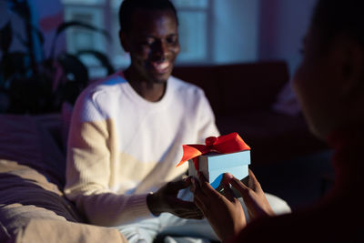 African man receives unexpected present from wife at home, woman giving gift box to happy boyfriend