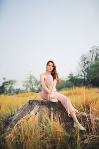 Portrait of young woman sitting on tree stump against clear sky