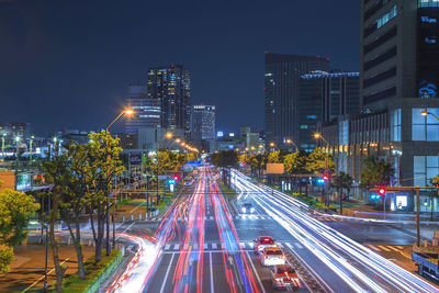 Light trails on city street by buildings against sky at night