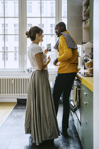 Full length of smiling woman holding smart phone talking with boyfriend doing chores in kitchen at home