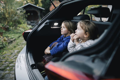 Girl day dreaming by sister while sitting in electric car trunk