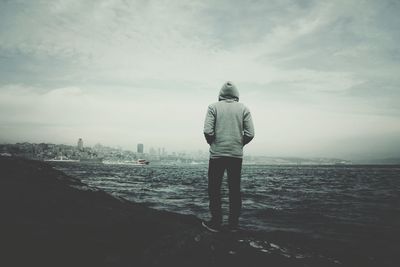 Rear view of man standing at shore against cloudy sky