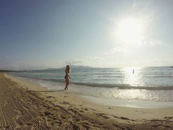 Side view of young woman in bikini standing at beach against sky during sunny day