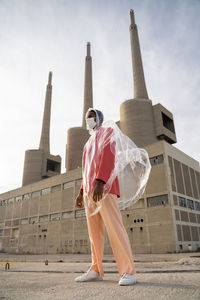 Man wearing raincoat and protective face mask looking away while standing against building