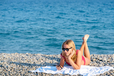 Woman relaxing laying on the pebble beach wearing pink bathing suit using her mobile smart phone